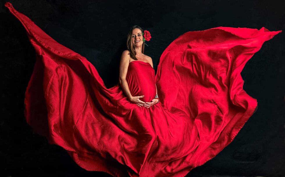 Mujer embarazada sonriente con una flor roja detrás de la oreja y cubierta con una tela roja que le marca la barriga
