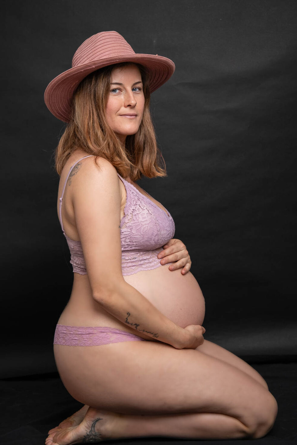 Mujer embarazada de perfil con lencería mirando a cámara sonriente en un estudio de fotografía