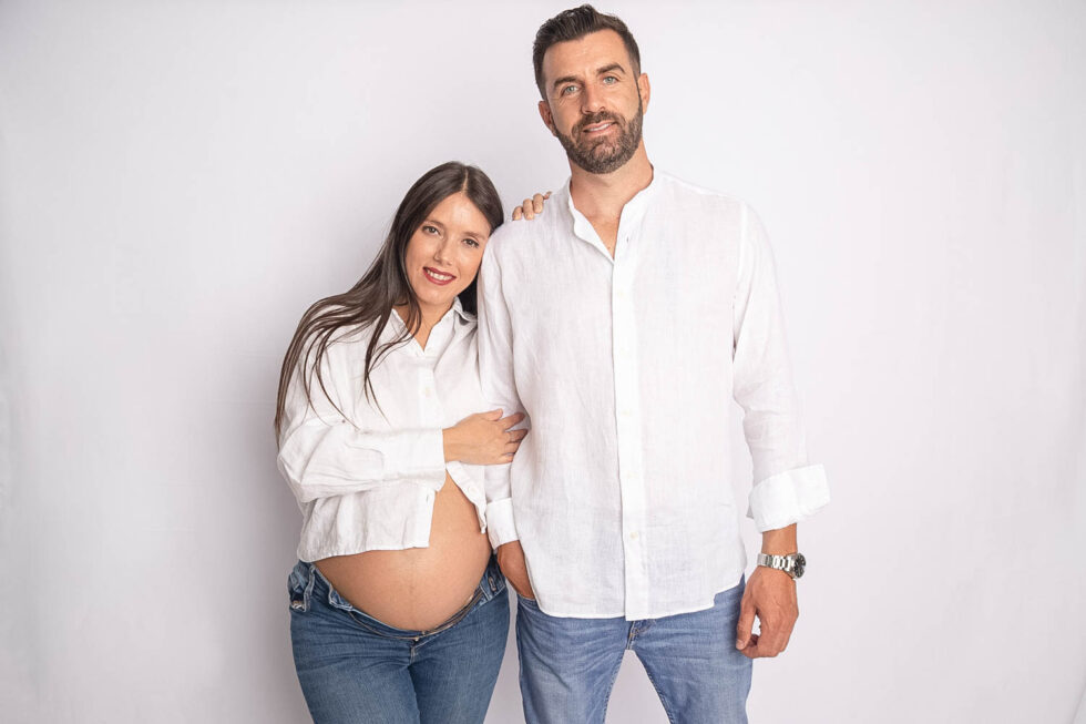 Mujer embarazada agarrada a su pareja, vestidos similares con vaqueros y camisa blanca miran sonrientes a la cámara