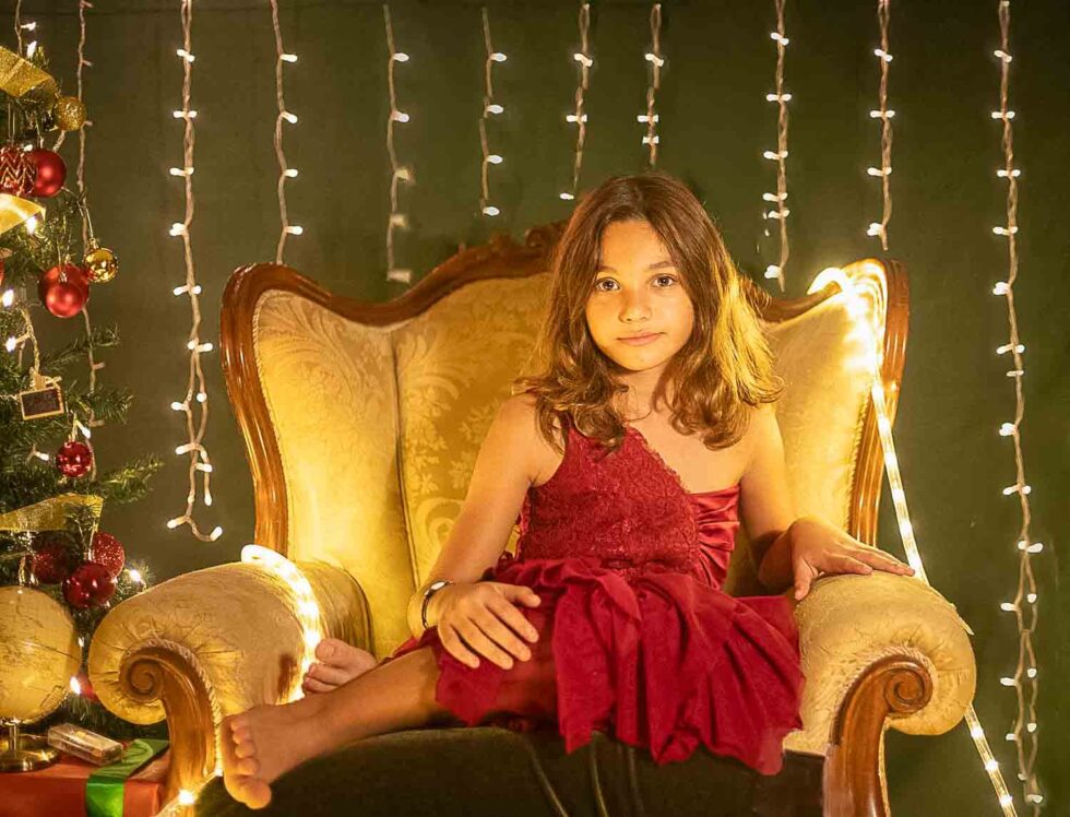 Niña vestida de rojo sentada en una bonita butaca con fondo de luces y con un árbol de Navidad al lado