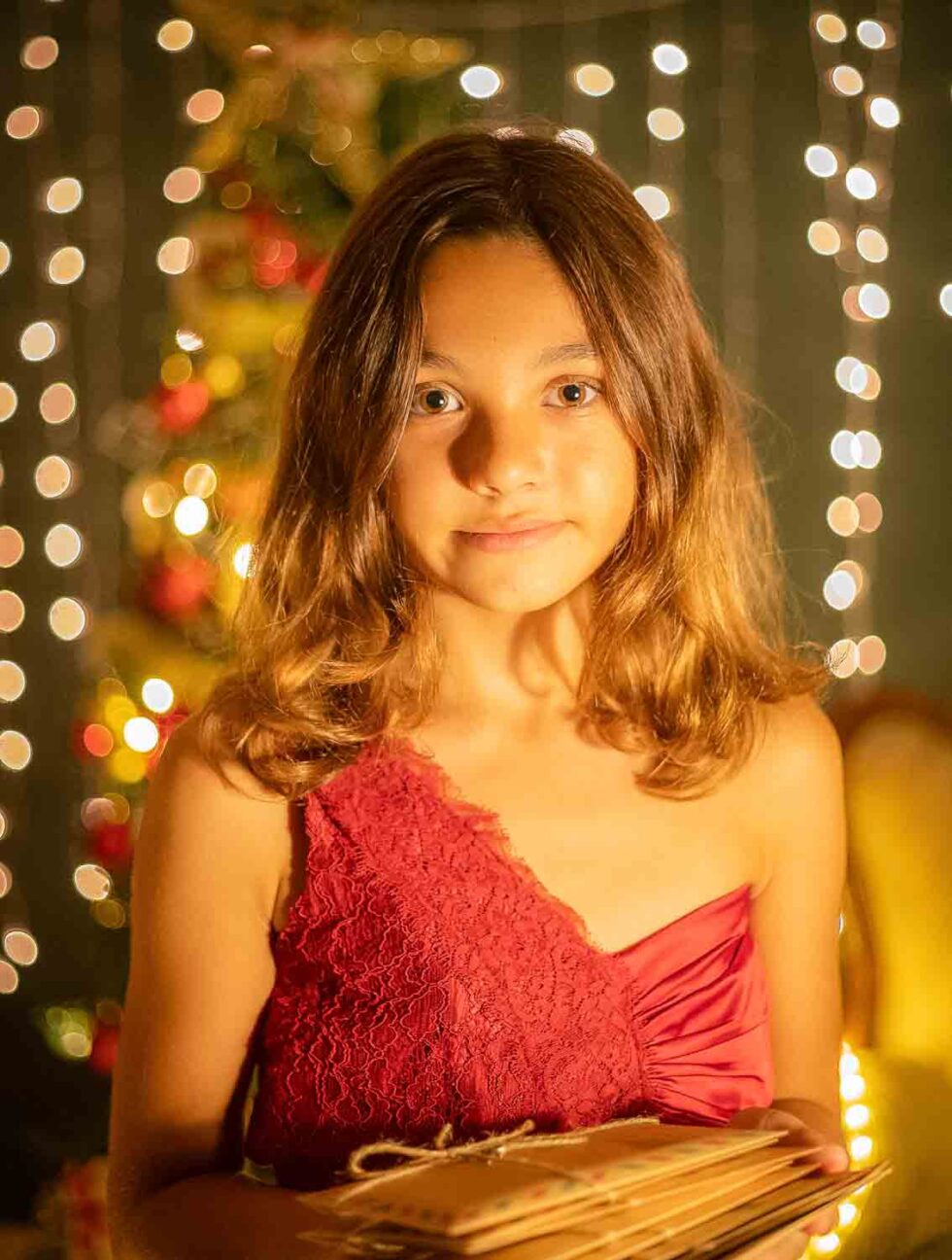 Niña mirando a cámara sosteniendo cartas de Navidad con un fondo navideño de luces y árbol