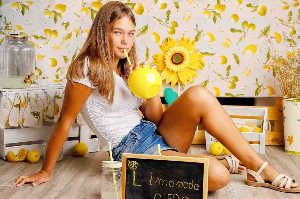 Chica sentada, bebiendo limonada y mirando a cámara sonriente en un bonito estudio de fotografía decorado con limones