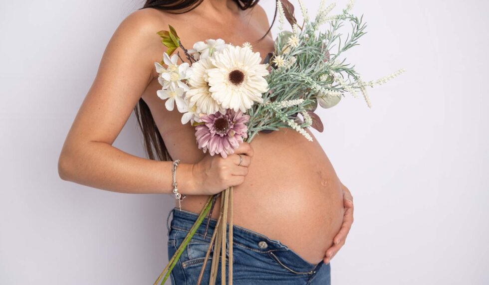 Plano medio de embarazada de perfil acariciándose la barriga con una mano y con la otra sujetando un ramo de flores que le cubre el pecho