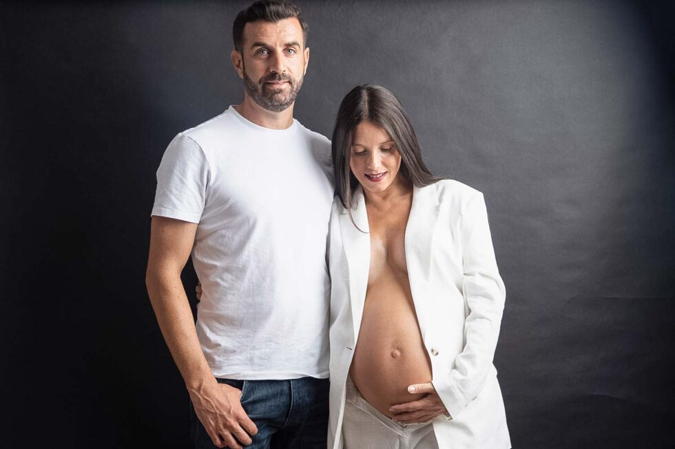 Fotografía de Mujer embarazada y su pareja vestidos de blanco con un fondo negro en el estudio de fotos