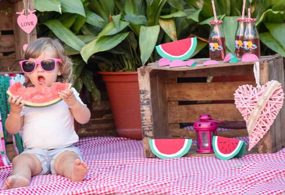 Niña pequeña con gafas rosas, sentada y boca abierta comiendo una rodaja de sandía con un fondo decorado con sandías muy divertido