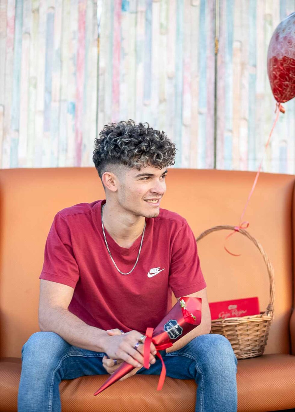 Chico sentado en un sofá con una flor en la mano junto a una cesta con bombones y un globo
