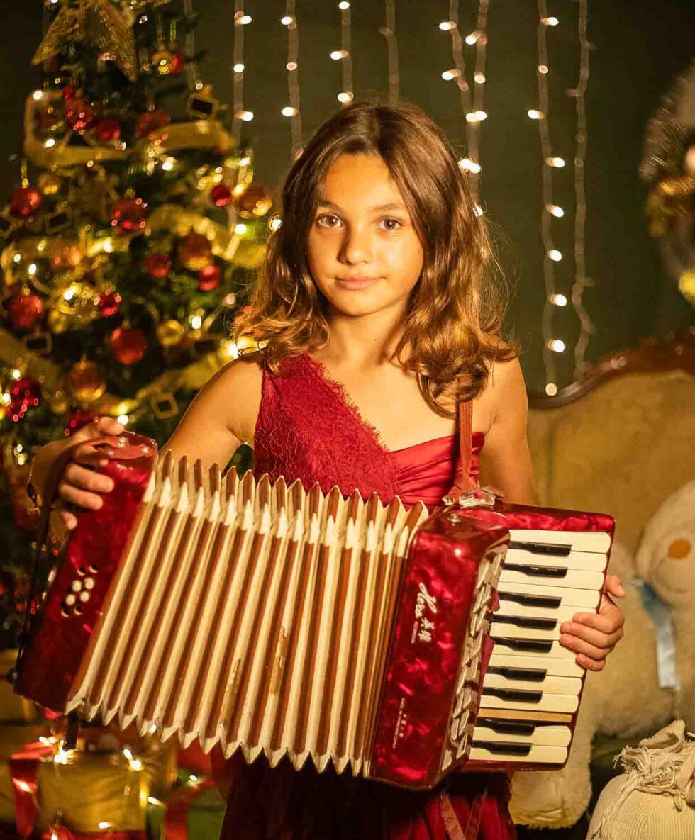 Chica vestida de rojo tocando el acordeón con un fondo navideño detrás con árbol y luces