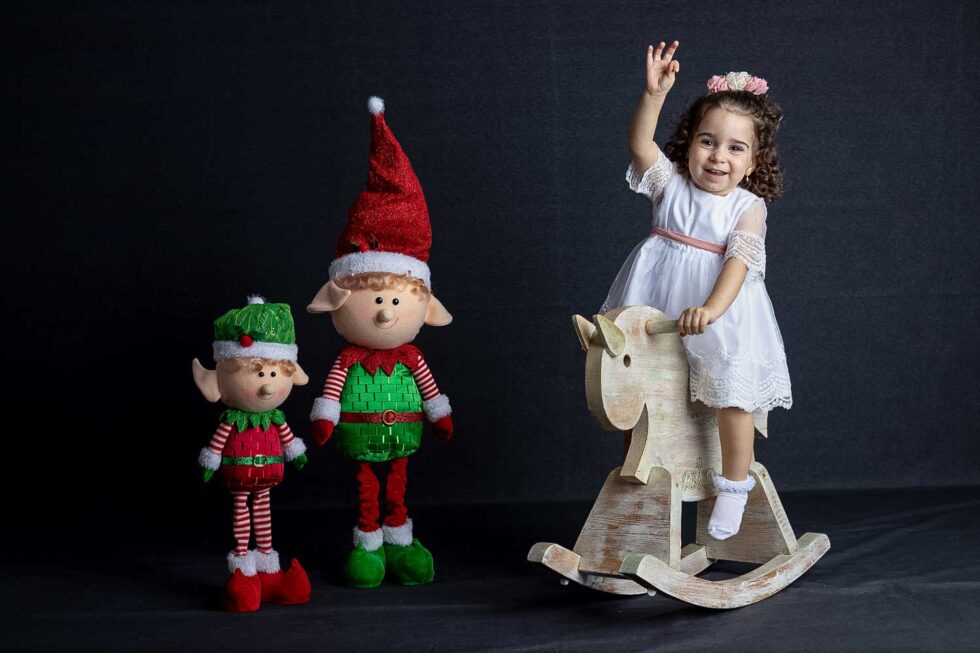 Niña pequeña saludando mientras se balancea en un caballito de madera con elfos decorativos al lado