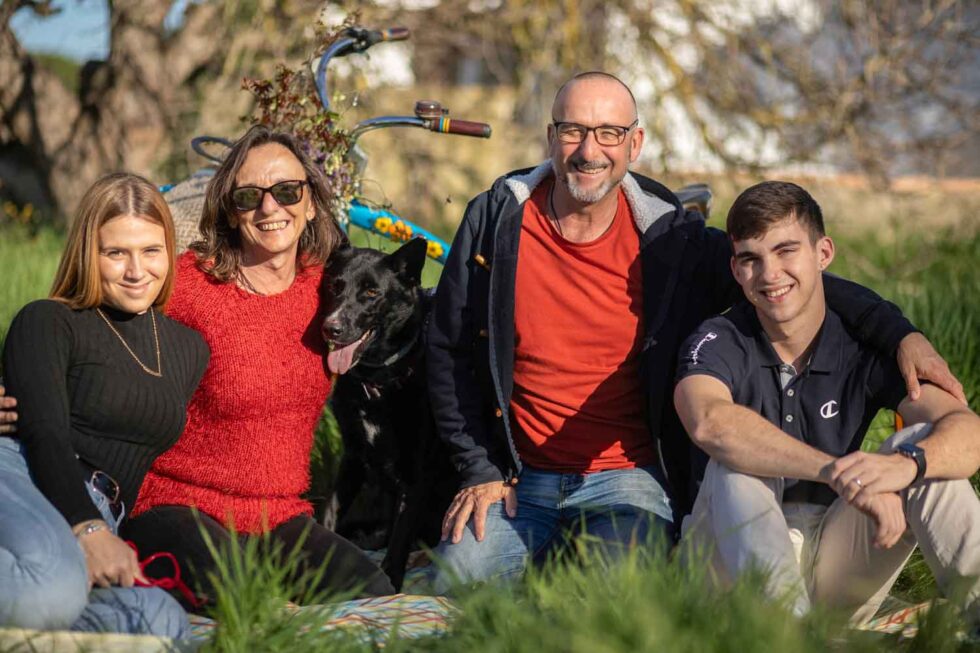 Familia de cuatro miembros y un perro sentados sobre la hierba muy sonrientes y mirando a cámara