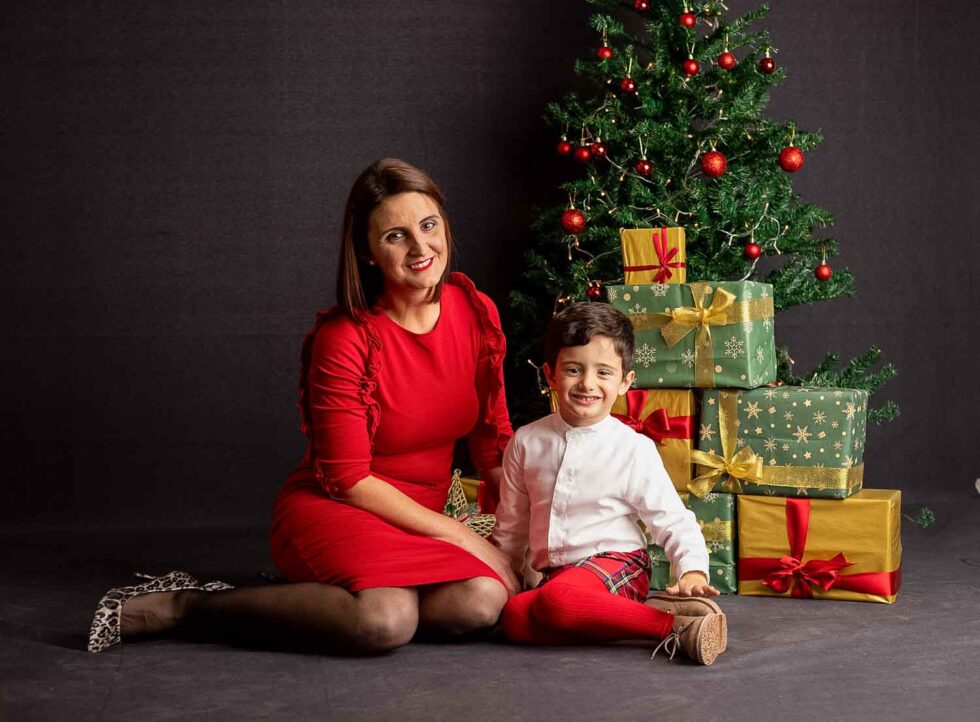 Madre e hijo vestidos de rojo posando en un decorado de fotografía con árbol y regalos navideños